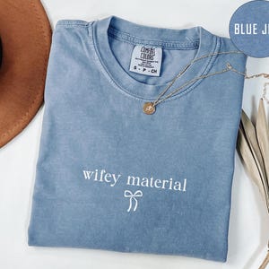 May include: A blue jean Comfort Colors t-shirt with the phrase "wifey material" printed in white. A gold necklace with a pendant is draped across the shirt. The shirt is folded and displayed on a white surface.