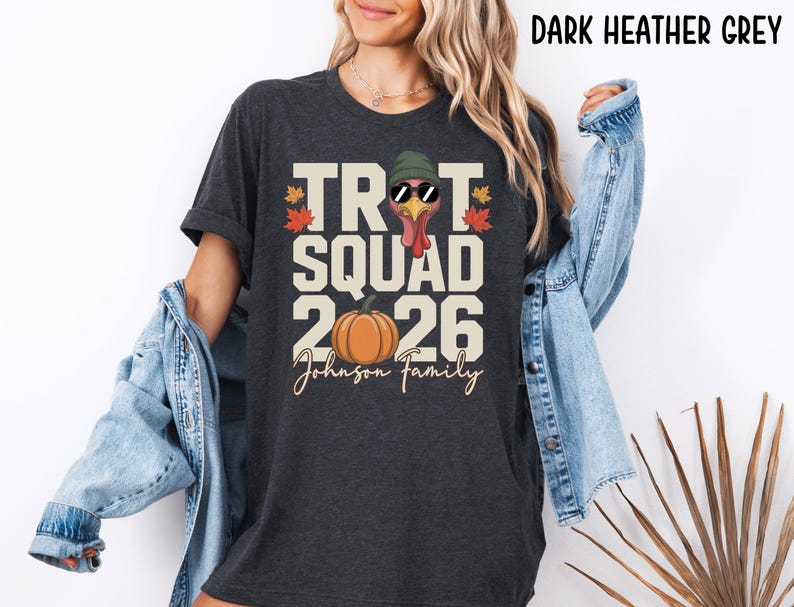 May include: A dark heather grey t-shirt featuring a graphic print with the text "TROT SQUAD 2026" and "Johnson Family". The design includes a turkey wearing a green beanie and sunglasses, an orange pumpkin, and small red and orange autumn leaves.