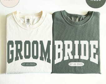 May include: Two t-shirts, one ivory and one moss green, are displayed. The ivory shirt says "GROOM" in green block letters, and the green shirt says "BRIDE" in white block letters. Both shirts have the date "07.10.2025" printed below the text.