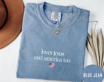 May include: A blue jean Comfort Colors t-shirt with the text "loves jesus and america too" and a heart-shaped American flag. A gold necklace with a pendant is draped across the shirt. A brown hat is visible in the upper left corner.