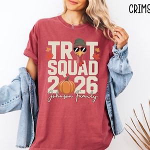 May include: A crimson short-sleeve t-shirt featuring a graphic of a turkey wearing a green beanie and sunglasses, with the text "TROT SQUAD 2026" and "Johnson Family" printed in cream, alongside a pumpkin and autumn leaves in orange and red.