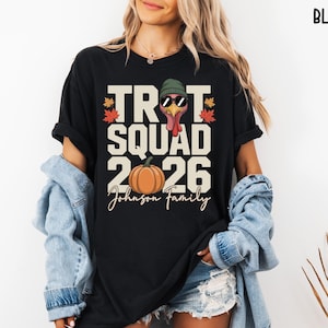May include: A black t-shirt featuring a graphic print with the text TROT SQUAD 2026 and Johnson Family. The design includes a turkey wearing a green beanie and sunglasses, an orange pumpkin, and colorful autumn leaves in red and orange tones.