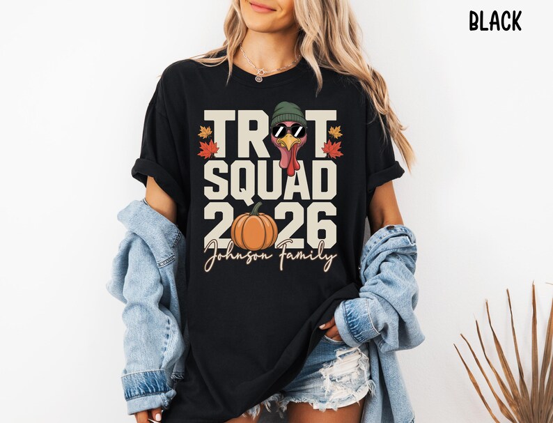 May include: A black t-shirt featuring a graphic print with the text TROT SQUAD 2026 and Johnson Family. The design includes a turkey wearing a green beanie and sunglasses, an orange pumpkin, and colorful autumn leaves in red and orange tones.