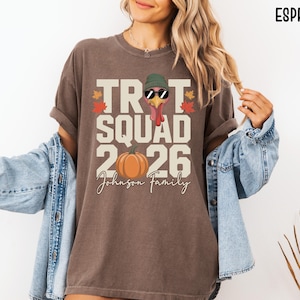 May include: An espresso-colored t-shirt featuring a graphic with the text TROT SQUAD 2026 and Johnson Family. The design includes a turkey wearing a green beanie and sunglasses, an orange pumpkin, and autumn leaves in red and yellow colors.