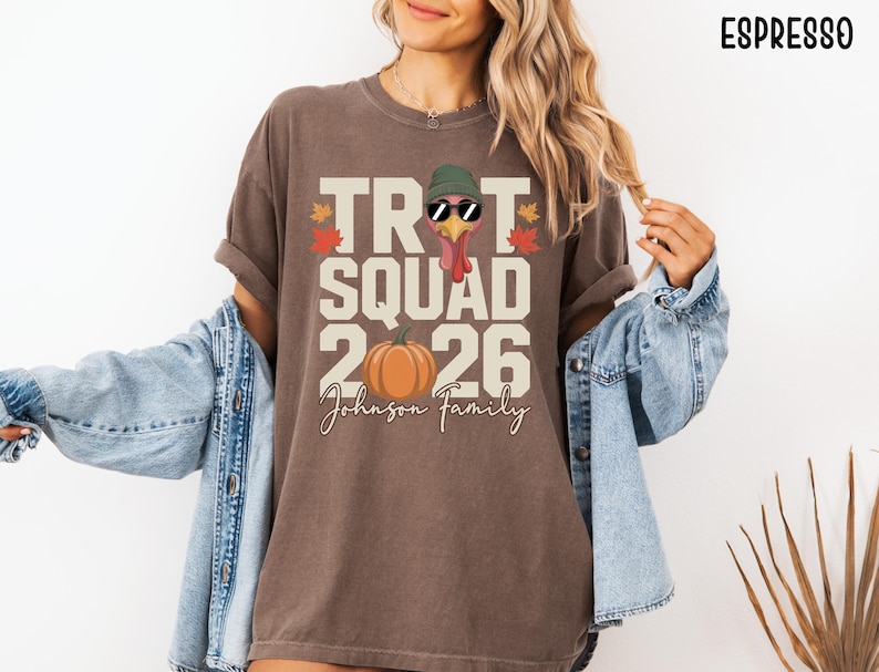 May include: An espresso-colored t-shirt featuring a graphic with the text TROT SQUAD 2026 and Johnson Family. The design includes a turkey wearing a green beanie and sunglasses, an orange pumpkin, and autumn leaves in red and yellow colors.