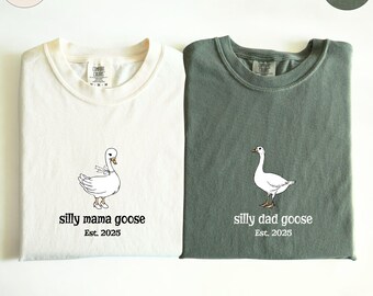 May include: Two folded t-shirts, one ivory and one moss green, with goose illustrations. The ivory shirt says "silly mama goose Est. 2025." The green shirt says "silly dad goose Est. 2025." Both shirts have a crew neck.