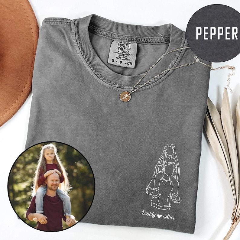 May include: A gray t-shirt with a white outline drawing of a father carrying a child on his shoulders. The text "Daddy &hearts; Alice" is below the drawing. A small photo of a father and child is in the lower left corner of the image.