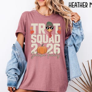 May include: A heather mauve t-shirt featuring a graphic with the text TROT SQUAD 2026 and Johnson Family. The design includes a turkey wearing a green beanie and sunglasses, an orange pumpkin, and small autumn leaves in red and orange colors.