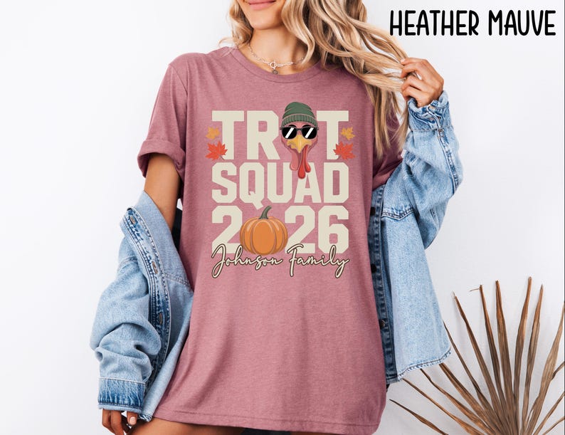 May include: A heather mauve t-shirt featuring a graphic with the text TROT SQUAD 2026 and Johnson Family. The design includes a turkey wearing a green beanie and sunglasses, an orange pumpkin, and small autumn leaves in red and orange colors.