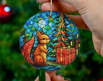 May include: A round Christmas ornament featuring a stained-glass design with a squirrel, Christmas tree, and gift box. The ornament has a gold string for hanging. The background is a blurred Christmas tree with red ornaments.