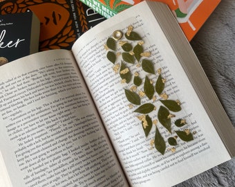 May include: A clear bookmark with a gold ring at the top. The bookmark is decorated with pressed green leaves and gold accents.