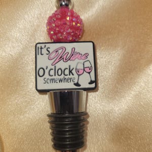 May include: Pink and white wine bottle stopper with the text "It's Wine o'clock Somewhere" and two wine glasses. The stopper has a silver top and a black bottom.