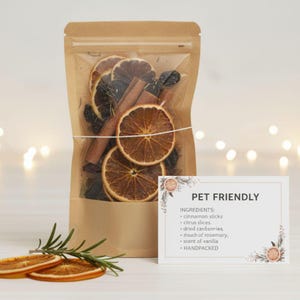 May include: A brown paper bag with a clear front, filled with dried orange slices, cinnamon sticks, and other ingredients. A white card with the text "PET FRIENDLY" and ingredient list is next to the bag. The ingredients include cinnamon sticks, citrus slices, and dried cranberries.
