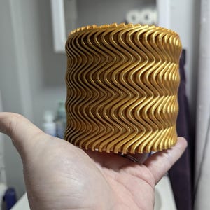 May include: A cylindrical object with a metallic gold finish and a repeating vertical wavy texture. The surface features a series of parallel, undulating ridges that wrap around the entire circumference of the item, creating a consistent, rhythmic pattern from top to bottom.