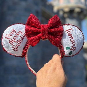May include: White mouse ears with red sequin bows and a red embroidered message that says "Very Merry Birthday" on one ear and "Tamara" with a holly sprig on the other ear.