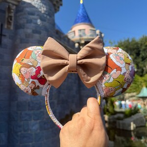 May include: A pair of brown Minnie Mouse ears with a brown bow. The ears are covered in a print of the seven dwarfs from Snow White and the Seven Dwarfs.