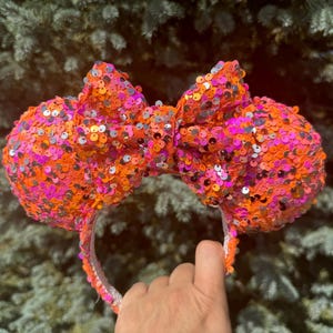May include: A pair of pink, orange, and silver sequined Minnie Mouse ears with a bow.