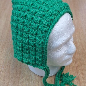 May include: A handmade, bright green crochet hat with a pointed top and ear flaps. The hat features a textured stitch pattern and tassels at the end of the ear flaps. The hat is displayed on a white mannequin head.