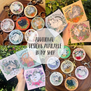 May include: A collection of colorful cat-themed keychains with different names, including Biscuit, Oliver, Hannah, Tammy, Lily, Thomas, Phoebe, and Sannah. The keychains feature illustrations of cats in various poses and settings, with some including flowers, hearts, and other decorative elements. The text "ADDITIONAL DESIGNS AVAILABLE IN MY SHOP" is displayed in the center of the image.