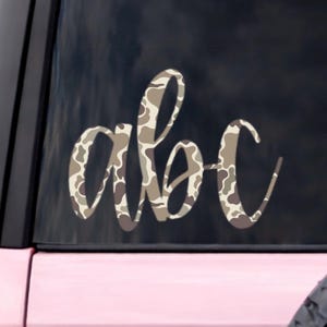 May include: A decorative car decal featuring the letters "abc" in a stylish, cursive font. The letters are filled with a camouflage pattern in shades of brown, tan, and cream. The decal is applied to the rear window of a pink vehicle.