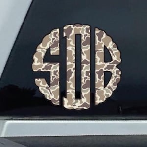 Pode incluir: Um autocolante circular para carro com as letras "SOB" num padrão de camuflagem. O autocolante está fixado no vidro traseiro de um veículo branco. O design de camuflagem apresenta tons de castanho e bege.