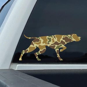 May include: A camouflage-patterned dog decal is affixed to the rear window of a white pickup truck. The dog is depicted in a walking pose, with a brown, tan, and green camouflage design. The background shows a blue sky and green foliage.