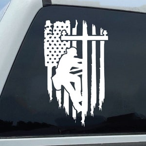 May include: White vinyl decal of a silhouette of a lineman climbing a utility pole with a cross on top, all in front of an American flag. The decal is on the back window of a white pickup truck.