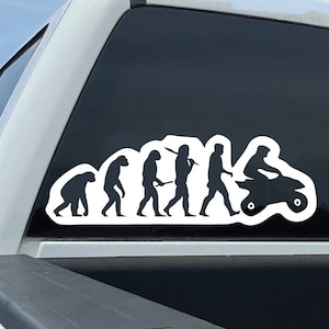 May include: A white truck with a black silhouette sticker on the back window. The sticker shows the evolution of man, ending with a person riding an ATV.