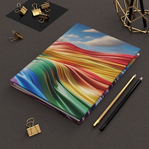 以下が含まれることがあります： 波打つデザインの虹色のノートブック。表紙は雲のある空。ノートブックは閉じられており、2本の鉛筆と金色のクリップと共に暗いグレーの表面の上に置かれている。