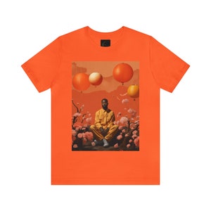 May include: Orange t-shirt with a graphic print of a person sitting in a field of flowers with orange and white lanterns floating in the sky.