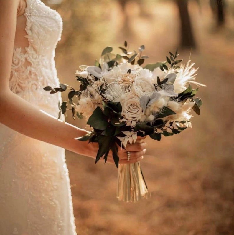 Pu&ograve; includere: Un bouquet da sposa con rose color crema, fiori secchi bianchi e fogliame verde scuro. La composizione &egrave; legata alla base con un nastro di colore chiaro, mostrando un mix di consistenze che include piume morbide e petali strutturati.