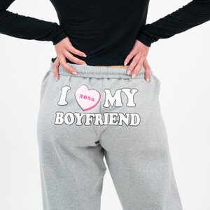 May include: Gray sweatpants with the text "I MY BOYFRIEND" in white block letters. A pink heart with "xoxo" is between the words "I" and "MY". The person is wearing a black long-sleeve shirt.