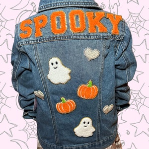 May include: A blue denim jacket with the word "SPOOKY" in orange felt letters on the back. The jacket is decorated with felt patches of ghosts, pumpkins, and hearts.