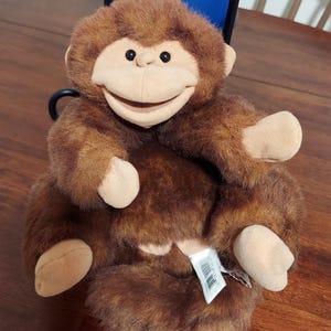 May include: A brown plush monkey puppet with a tan face, ears, hands, and feet. The monkey has black plastic eyes and a wide, smiling mouth. A white fabric tag with a barcode is attached to the lower body of the soft, furry toy.