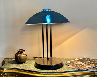 May include: A contemporary desk lamp with a dark teal, semi-circular shade and three black support rods. The lamp has a bright blue light and a round, black base. A small, bronze-coloured decorative vase is next to the lamp.