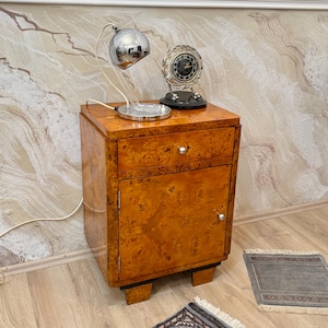 May include: A vintage wooden bedside cabinet with a burl wood finish. It has a drawer and a cabinet door with a silver knob. A chrome desk lamp and a decorative clock sit on top. The cabinet is on a wooden floor.