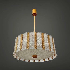 Könnte beinhalten: Ein runder Vintage-Kronleuchter mit strukturiertem Glasschirm und goldfarbenen Metallakzenten. Die Leuchte hängt an einer goldfarbenen Stange und Deckenhalterung. Das Design weist vertikale Goldleisten und einen dekorativen unteren Rand auf.