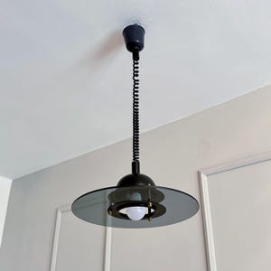 Könnte beinhalten: Eine schwarze und graue Pendelleuchte mit einem kreisförmigen, rauchfarbenen Glasschirm. Das Licht hängt an einem schwarzen, spiralförmigen Kabel und ist an der Decke befestigt. Die Leuchte hat ein modernes, minimalistisches Design.
