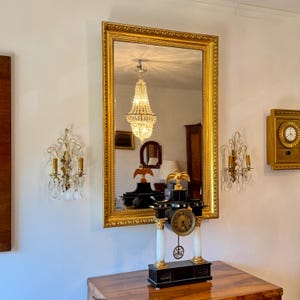 Puede incluir: Un espejo con marco dorado refleja una lámpara de araña y un reloj antiguo. El reloj es negro y blanco con detalles dorados, y está sobre una superficie de madera. Dos apliques de pared con cristales y velas flanquean el espejo.