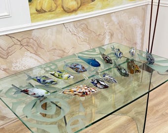 May include: A glass table with a clear top and a decorative etched design. The table displays a collection of colorful glass fish sculptures. The fish feature various patterns and colors, including blue, yellow, and red. The table is set against a backdrop of a wall painting.