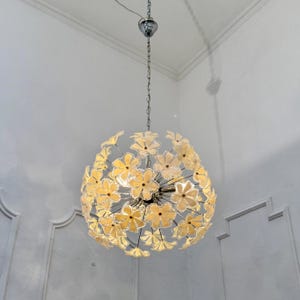 Op de afbeelding: Een bolvormige kroonluchter met een chromen afwerking en een veelvoud aan gele bloemvormige glazen kappen. De armatuur hangt aan een ketting en een chromen plafondbevestiging. Een decoratief item voor in huis.