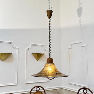 Könnte beinhalten: Eine Vintage-Hängeleuchte mit einem strukturierten, bronzefarbenen Schirm. Die Leuchte hat einen Holzsockel und ein Spiralkabel. Die Leuchte ist an einer Kette aufgehängt und leuchtet, wodurch ein warmes Licht entsteht. Die Leuchte ist ein dekoratives Element.