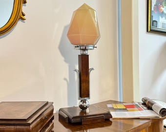 Peut inclure: Une lampe de table avec un abat-jour en verre facetté de couleur pêche pâle sur une colonne centrale en bois. La base est carrée et faite de bois foncé, agrémentée de détails en métal chromé poli, dont un élément sphérique au-dessus du socle.