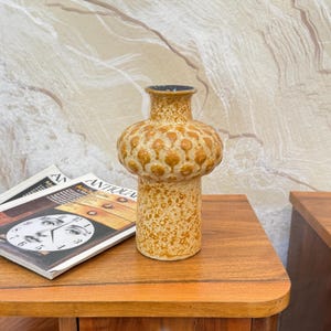 Op de afbeelding: Een vintage keramische vaas met een gespikkelde, bruine basis en een bolvormig, getextureerd lichaam. Het bovenste gedeelte heeft verhoogde, cirkelvormige ontwerpen in een donkerdere bruine tint. De vaas staat op een houten oppervlak.