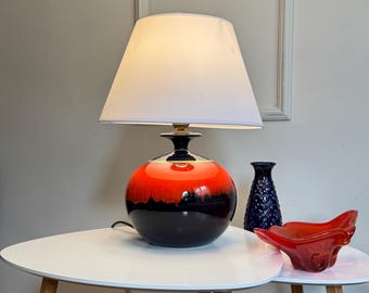 May include: A black and orange ceramic table lamp with a white lampshade. The lamp is on a white table with a red bowl.