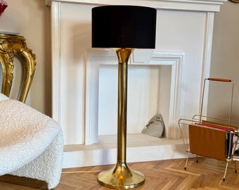 May include: A brass floor lamp with a black lampshade. The lamp has a round base and a slender stem. The lampshade is cylindrical. The lamp is in front of a white fireplace. The lamp's height is approximately 150 cm.