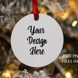 May include: A circular, white Christmas bauble with the words "Your Design Here" in black script. It is suspended by a red ribbon against a blurred backdrop of a Christmas tree with lights. The image also includes the text "PSD, PNG & JPG FILES INCLUDED".