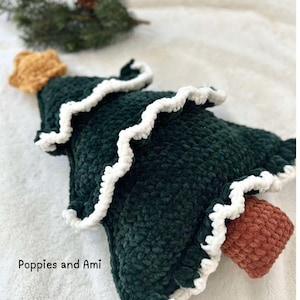 May include: A green crocheted Christmas tree cushion with a white crocheted trim and a brown crocheted star on top. The cushion is sitting on a white fluffy surface. "Poppies and Ami" is written in the bottom left corner of the image.