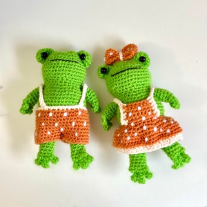 Op de afbeelding: Twee gehaakte kikker knuffels die bijpassende oranje en witte stippen outfits dragen. De kikker links draagt een overall en de kikker rechts draagt een jurk.