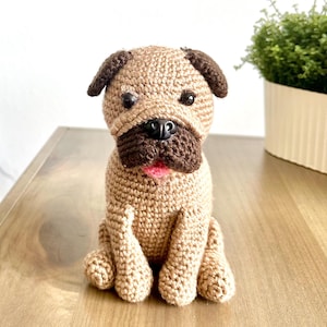 Può includere: Un giocattolo di cane carlino all'uncinetto con orecchie marroni e una lingua rosa. Il giocattolo è seduto su una superficie di legno.